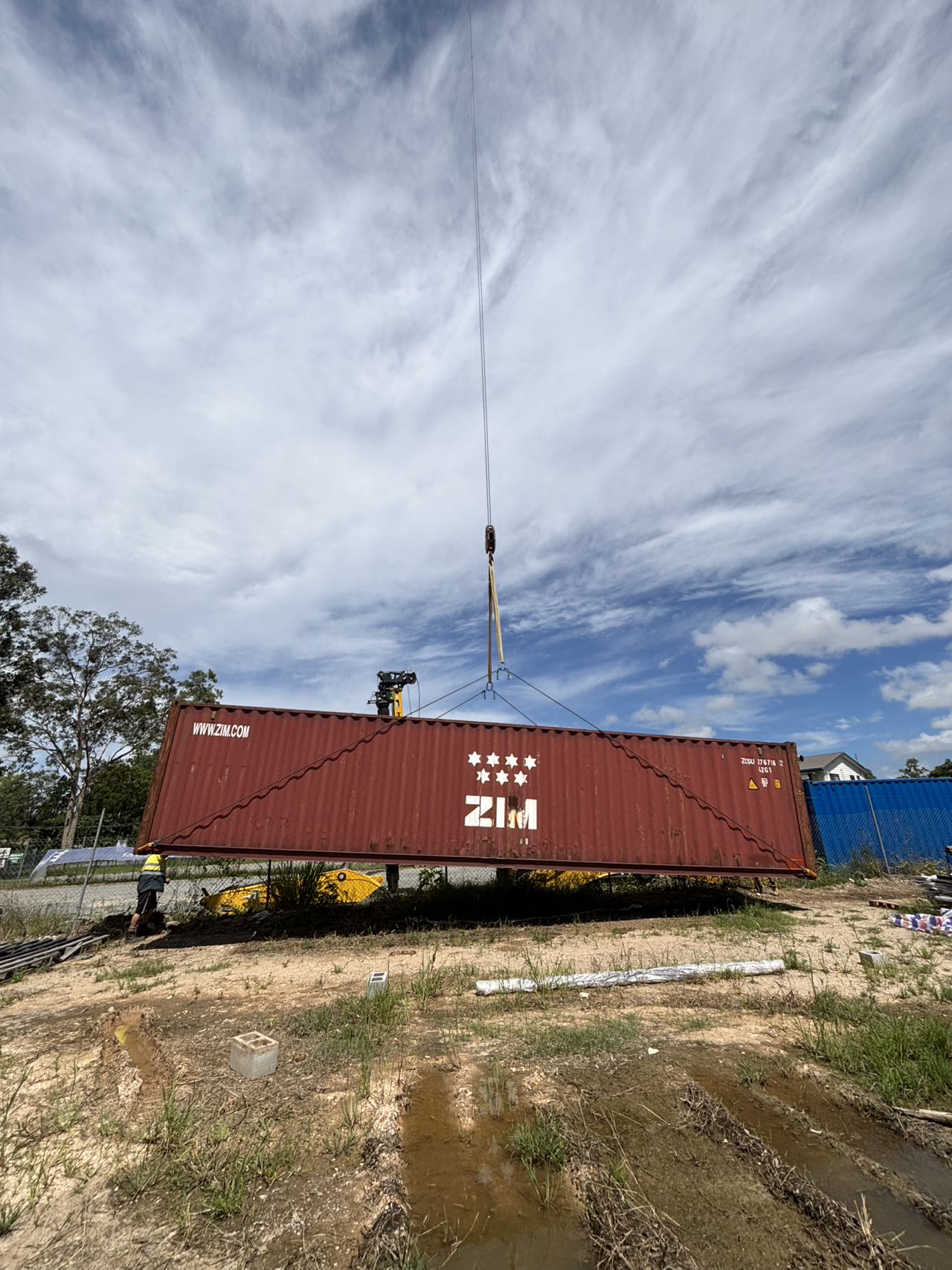 Red shipping container being lifted by a crane against a blue sky with clouds.