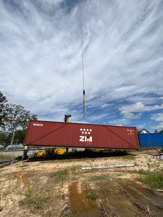 Red shipping container being lifted by a crane against a blue sky with clouds.