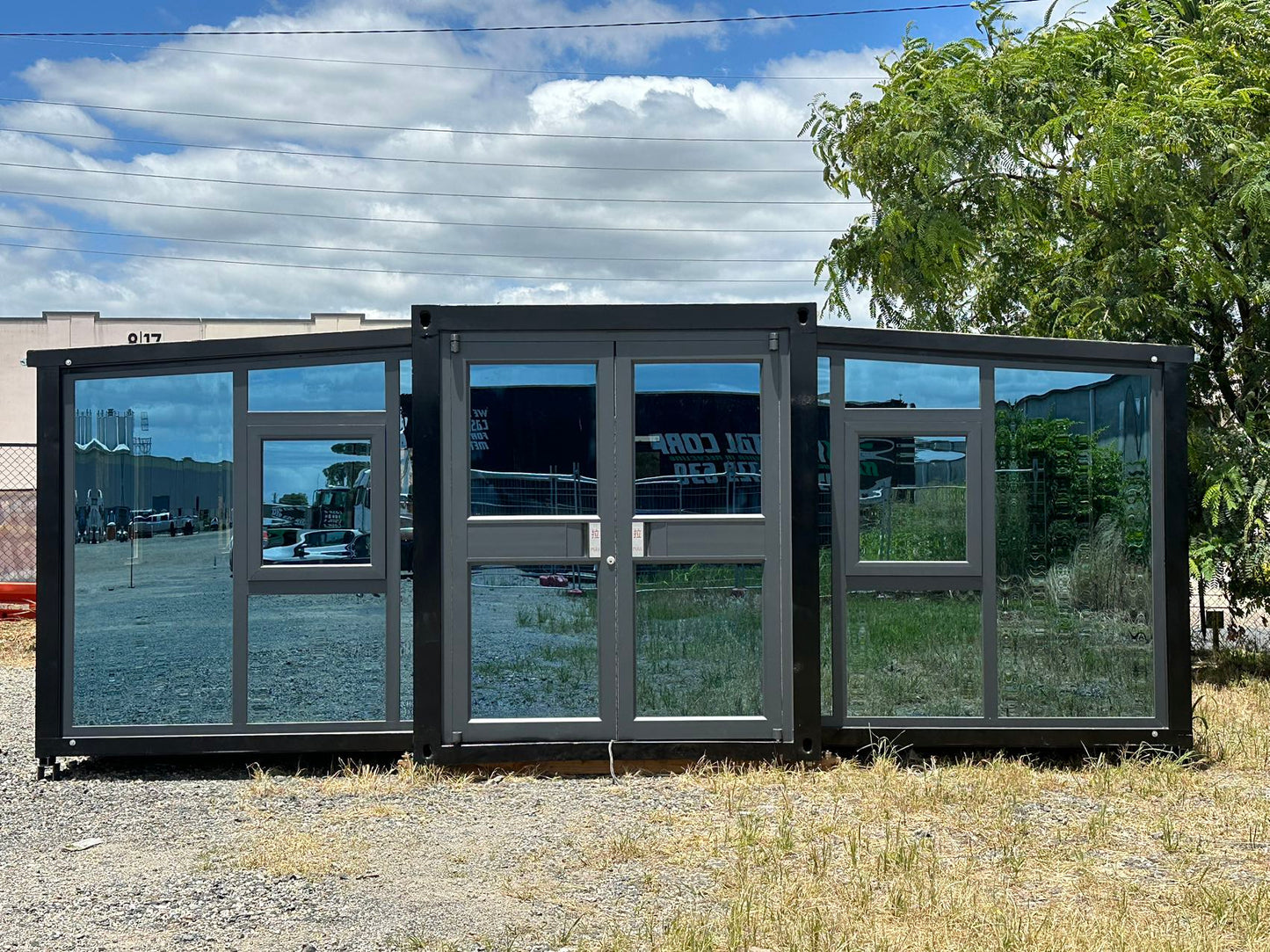 A front-faced glass curtain wall on a container house, with a clear blue sky and trees in the background.